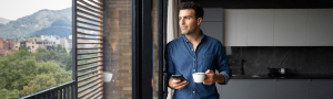 PIA Eine Person, steht in einer modernen Küche und hält eine Tasse Kaffee in der einen Hand und ein Smartphone in der anderen. Im Hintergrund sind große Fenster mit Blick auf eine Landschaft mit Bäumen und Bergen zu sehen.
